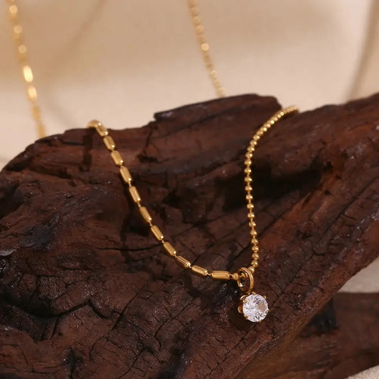 Elegant asymmetric bead chain necklace with a gold finish and a sparkling round pendant, displayed on a wooden surface. Perfect for stylish accessorizing.